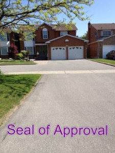 A driveway in need of a coat of sealant next to a groomed front lawn leading up to a two-story house with two garage doors. The words at the bottom read “Seal of Approval”